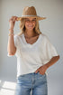 A woman wearing a white short-sleeve V-neck top, light blue jeans, and a straw hat, posing against a light gray background.