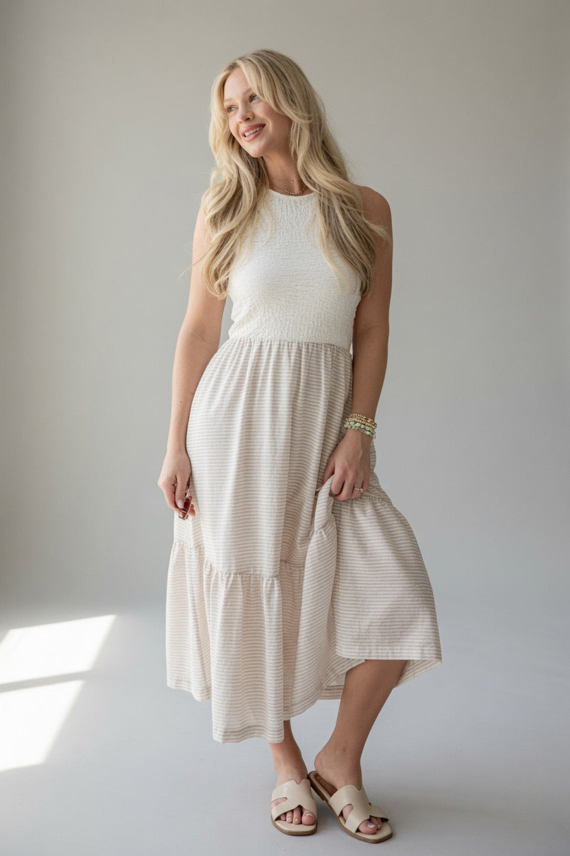A woman modeling a sleeveless tiered midi dress in a warm off-white or cream color, paired with neutral sandals, set against a plain light gray studio background.
