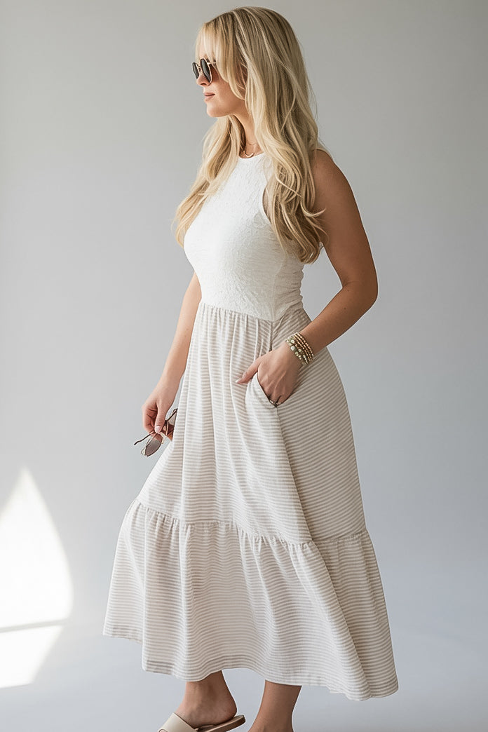 A woman modeling a sleeveless tiered midi dress with a textured cream bodice and fine horizontal stripes on the skirt, set against a plain light gray background.