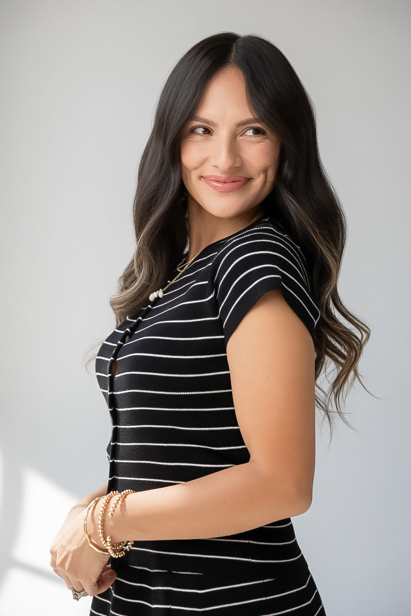 A woman with long dark brown hair is wearing a black short-sleeve top with thin white horizontal stripes, accessorized with gold bracelets, against a light gray studio background.