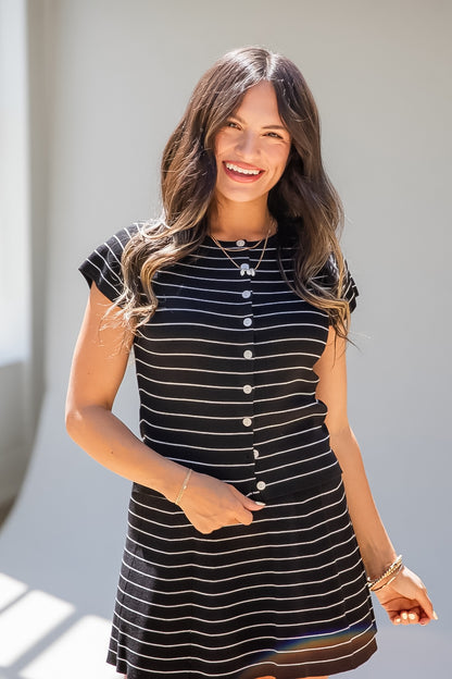 A woman wearing a black and white horizontally striped short-sleeve button-up dress, smiling in bright natural light against a neutral light gray wall.
