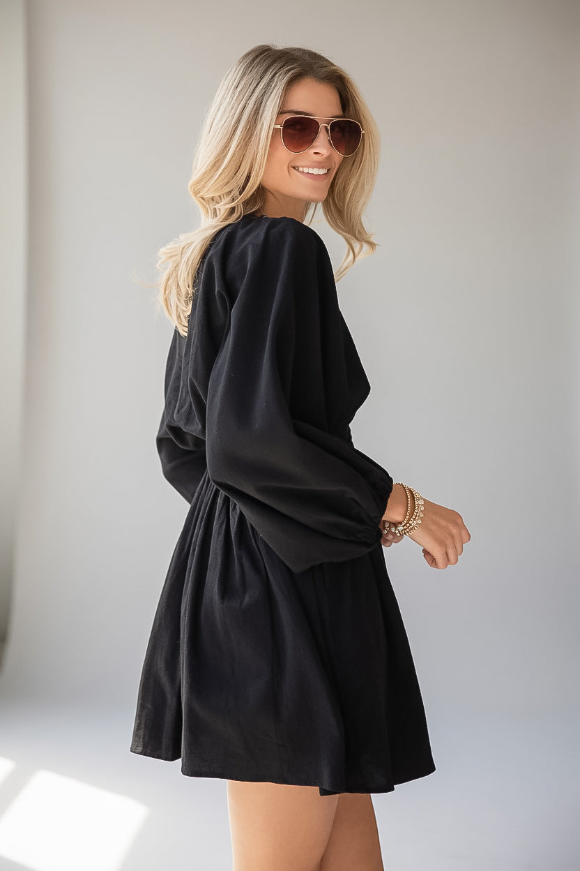 A woman modeling a black long-sleeve mini dress with a flared skirt, photographed in a bright indoor setting with a light gray background.