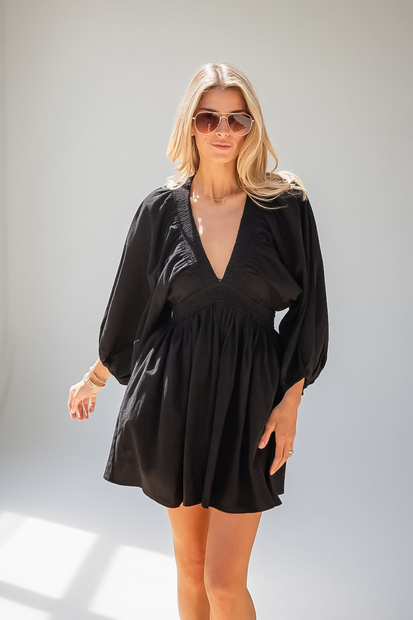 A blonde woman wearing a black long-sleeve mini dress with a deep V-neck and sunglasses, standing against a light gray studio background.