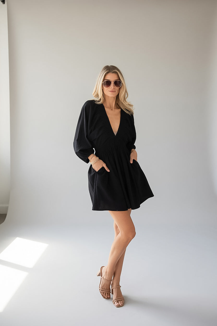 A woman modeling a black long-sleeve mini dress with a deep V-neck and side pockets, paired with nude high-heeled sandals and sunglasses, standing in a bright white studio.
