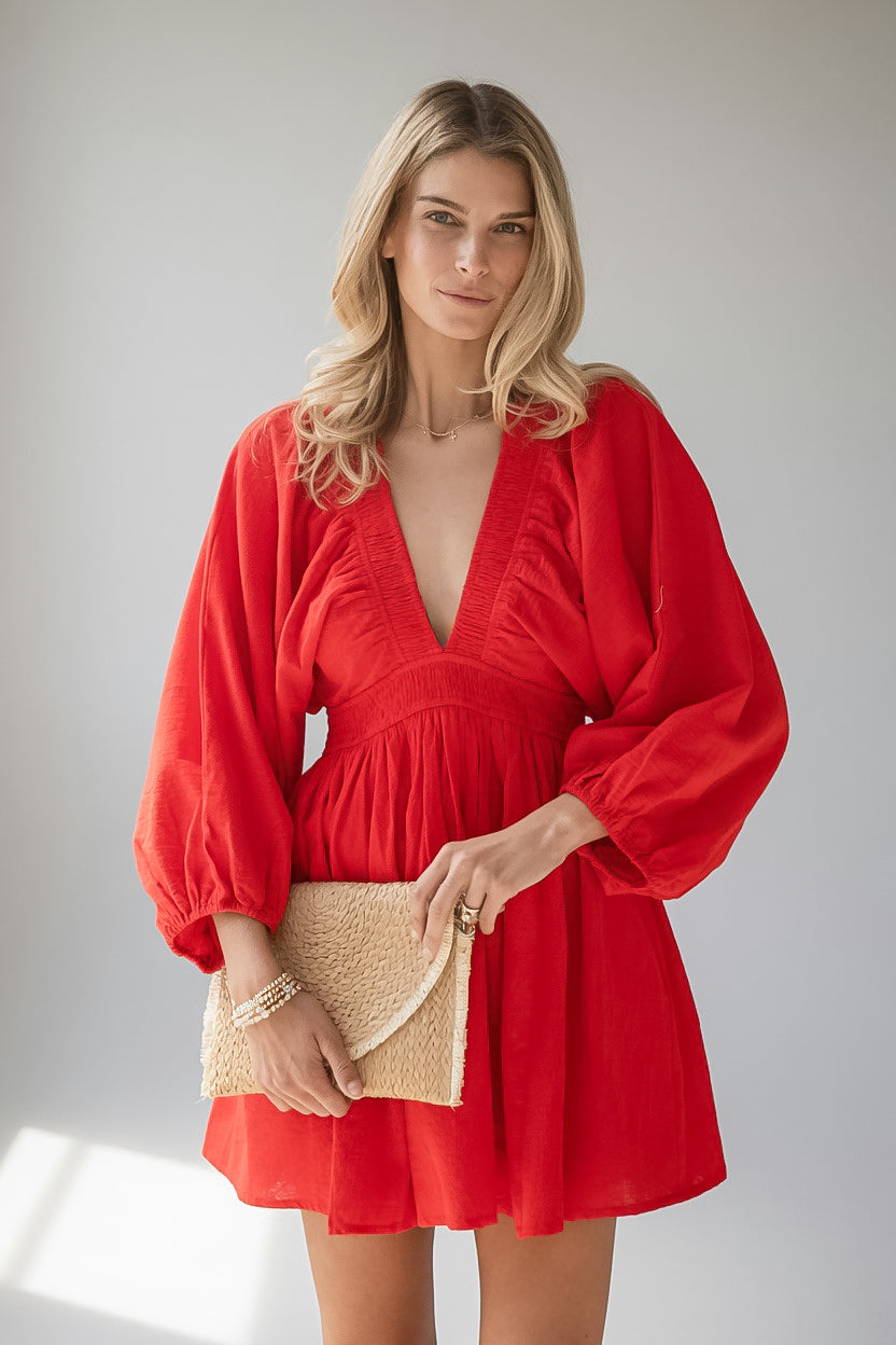 A woman modeling a bright red mini dress with voluminous balloon sleeves, a deep V-neckline, and a gathered waist, holding a woven beige clutch against a light gray studio background.