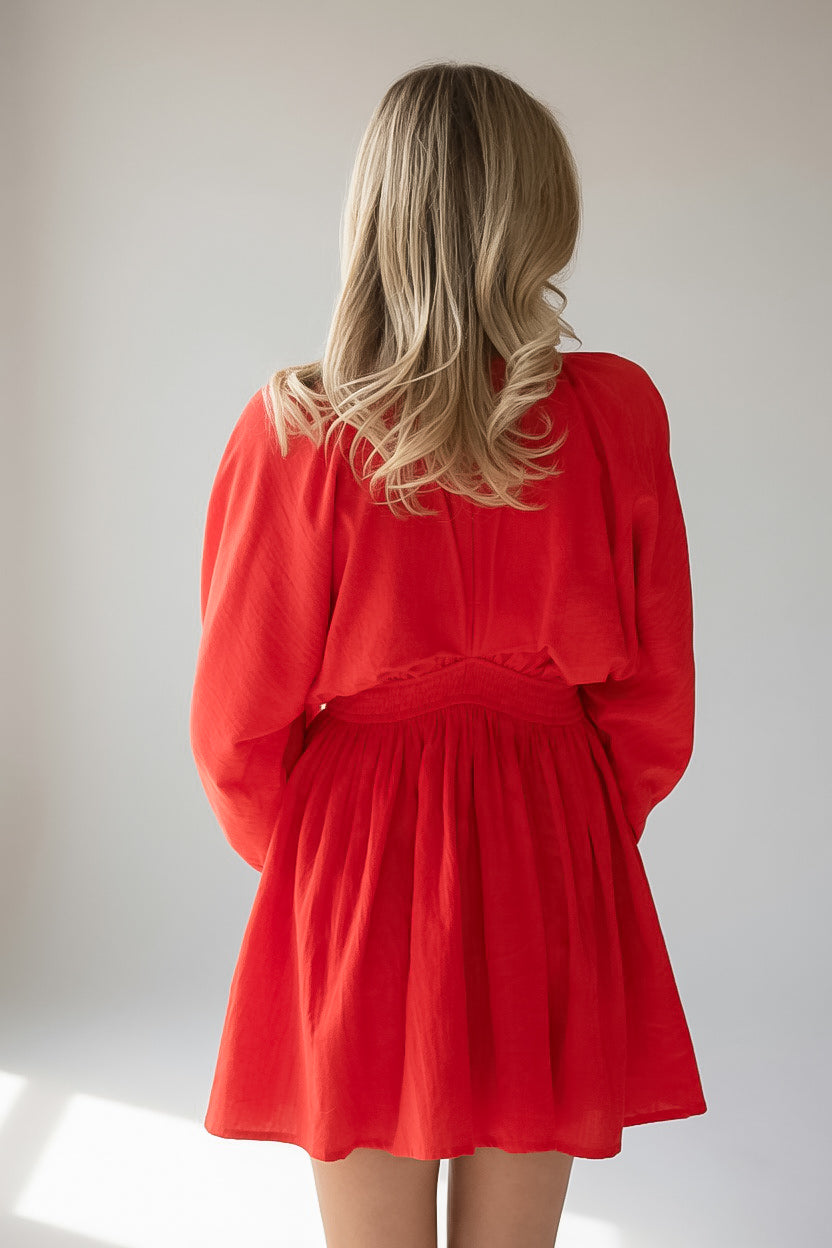 A woman with blonde hair is shown from behind wearing a bright red long-sleeve mini dress with a gathered waist, set against a plain light gray background.