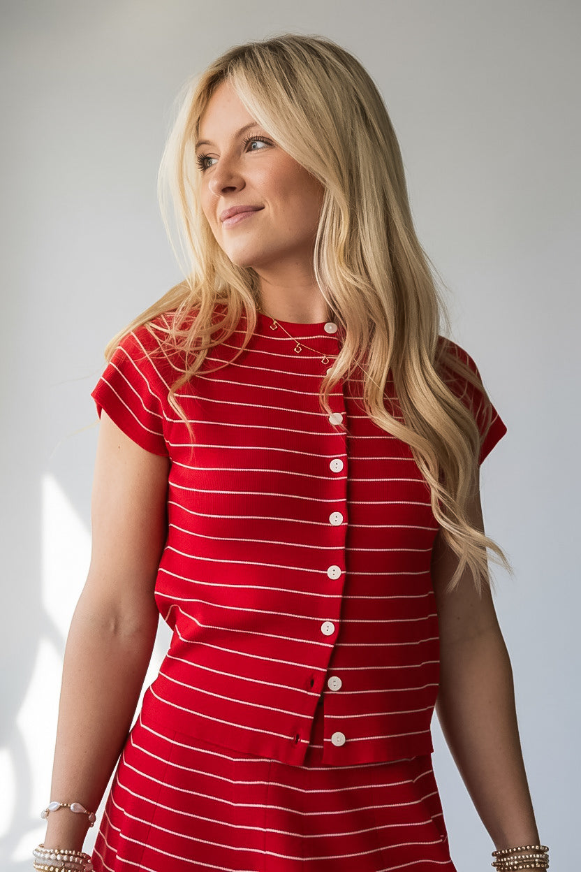 A blonde woman wearing a red short-sleeve top with thin white horizontal stripes and a button-down front, photographed against a light gray studio background.