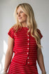 A blonde woman wearing a red short-sleeve top with thin white horizontal stripes and a button-down front, photographed against a light gray studio background.