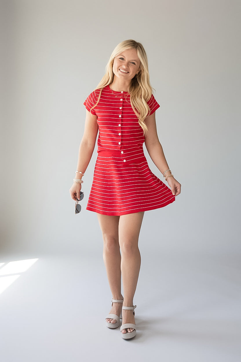 A young blonde woman modeling a red and white striped short-sleeve mini dress with a button-down front, standing against a light gray studio background.
