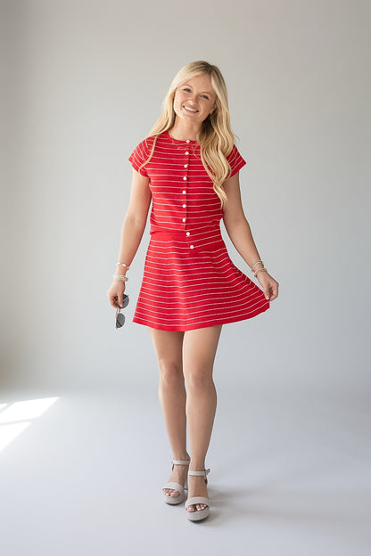 A young blonde woman modeling a red and white striped short-sleeve mini dress with a button-down front, standing against a light gray studio background.