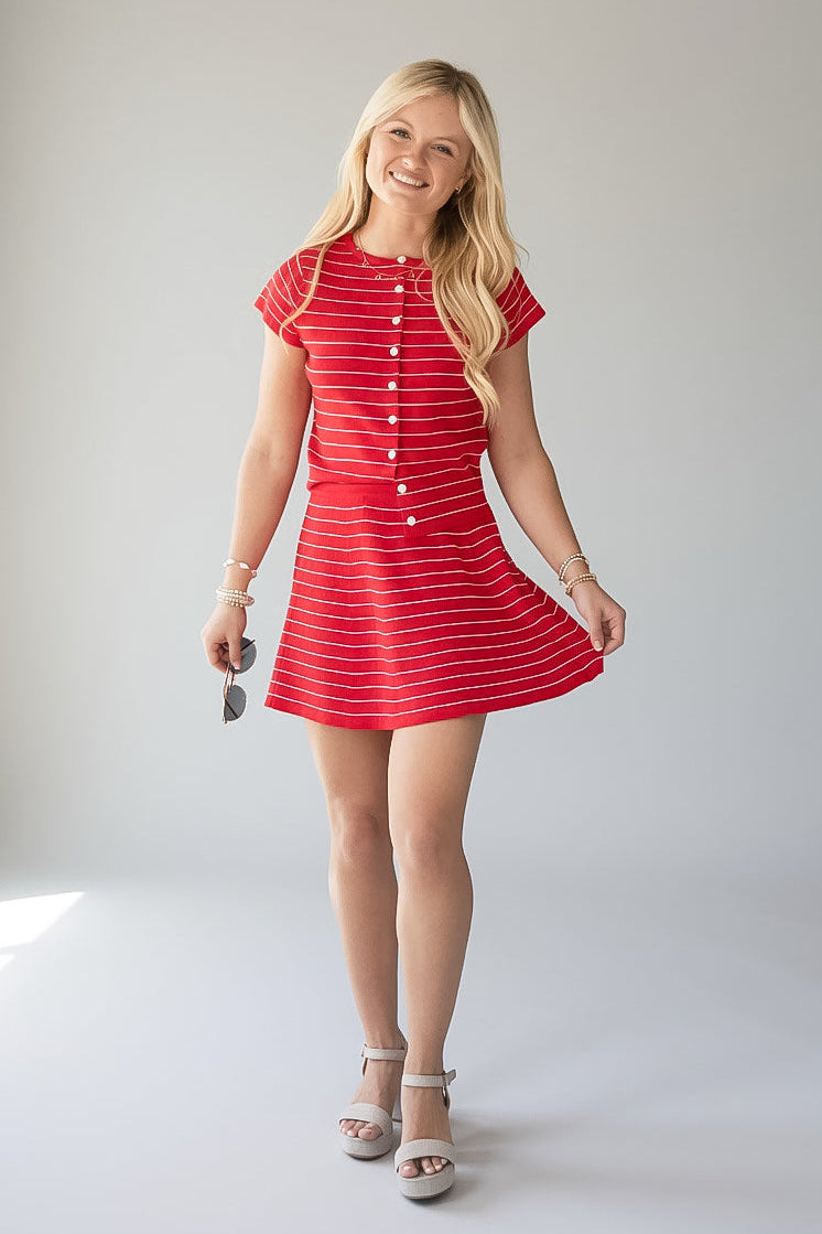 A young blonde woman modeling a red and white striped short-sleeve mini dress with a button-down front, standing against a light gray studio background.