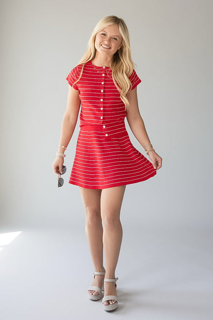 A young blonde woman modeling a red and white striped short-sleeve mini dress with a button-down front, standing against a light gray studio background.
