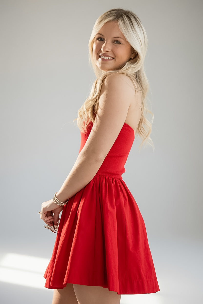 A blonde woman wearing a bright red strapless mini dress, posing in a studio setting against a light gray background.