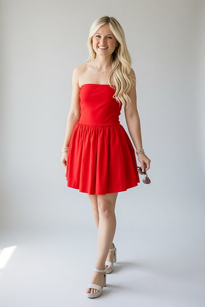 A smiling blonde woman models a strapless bright red mini dress with a flared skirt, paired with beige heeled sandals and holding a small purse, against a plain light gray studio background.