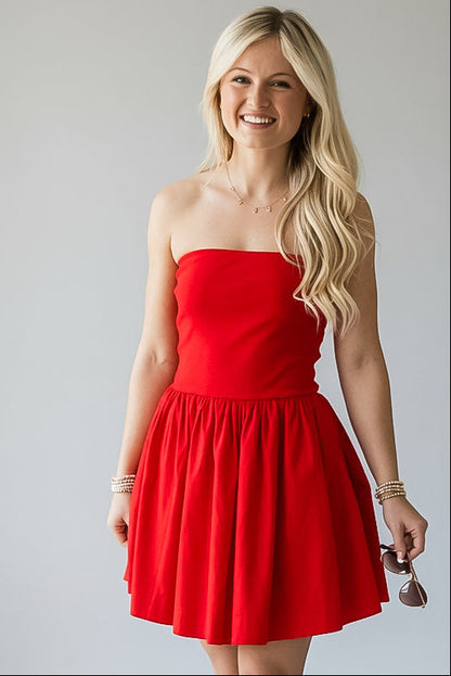 A smiling blonde woman wearing a bright red strapless mini dress with a flared skirt, standing against a light gray studio background and holding sunglasses.
