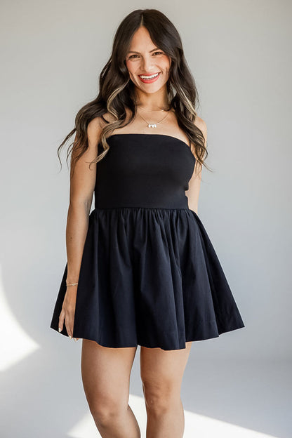 A woman modeling a strapless black mini dress with a flared skirt, standing against a light gray studio background with soft shadows.