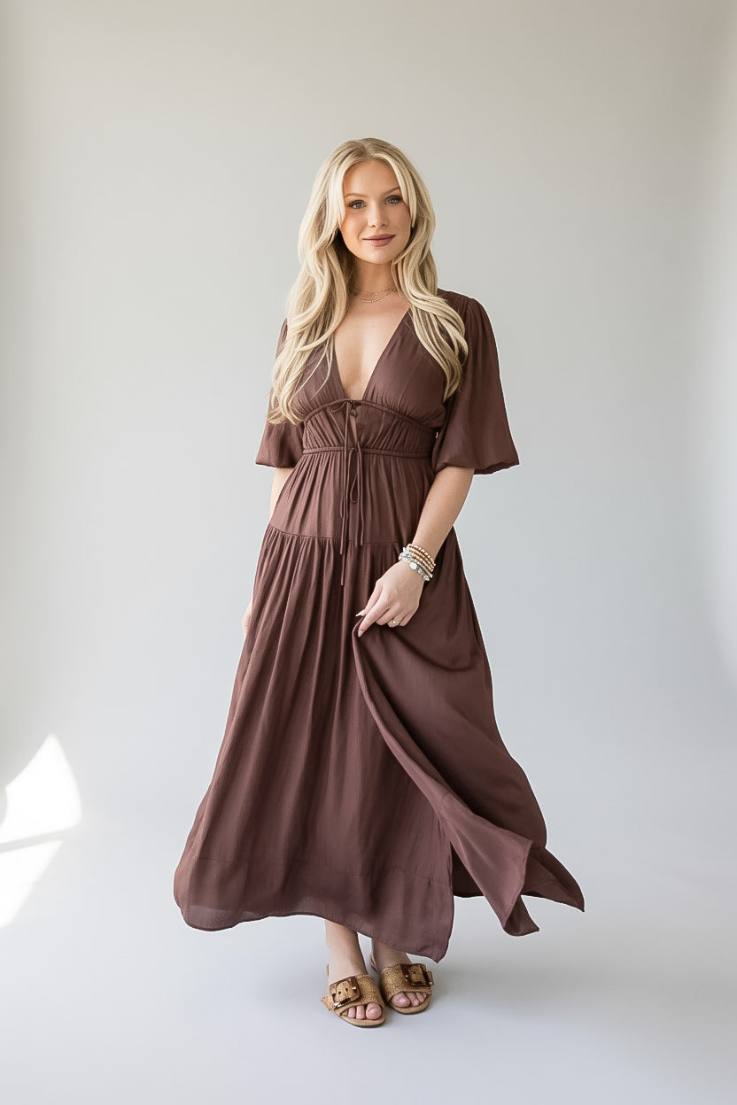 A blonde woman modeling a brown, knee-length maxi dress with short flutter sleeves, a deep V-neckline, and a gathered waist, standing against a neutral light gray studio background.