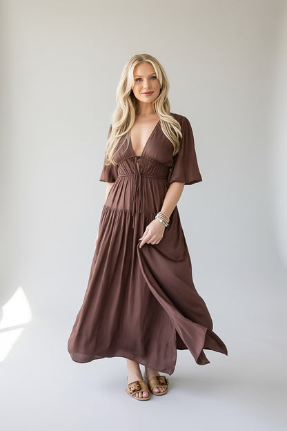 A blonde woman modeling a brown, knee-length maxi dress with short flutter sleeves, a deep V-neckline, and a gathered waist, standing against a neutral light gray studio background.