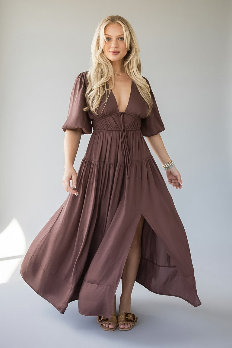 A blonde model wearing a dusty brown maxi dress with puff sleeves, a deep V-neckline, and a gathered waist, standing against a neutral light gray studio background.