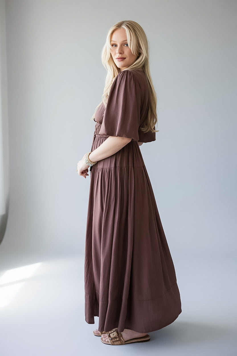 A woman modeling a long, dusty brown puff-sleeve maxi dress with a gathered waist, standing in a bright, minimalist studio setting.