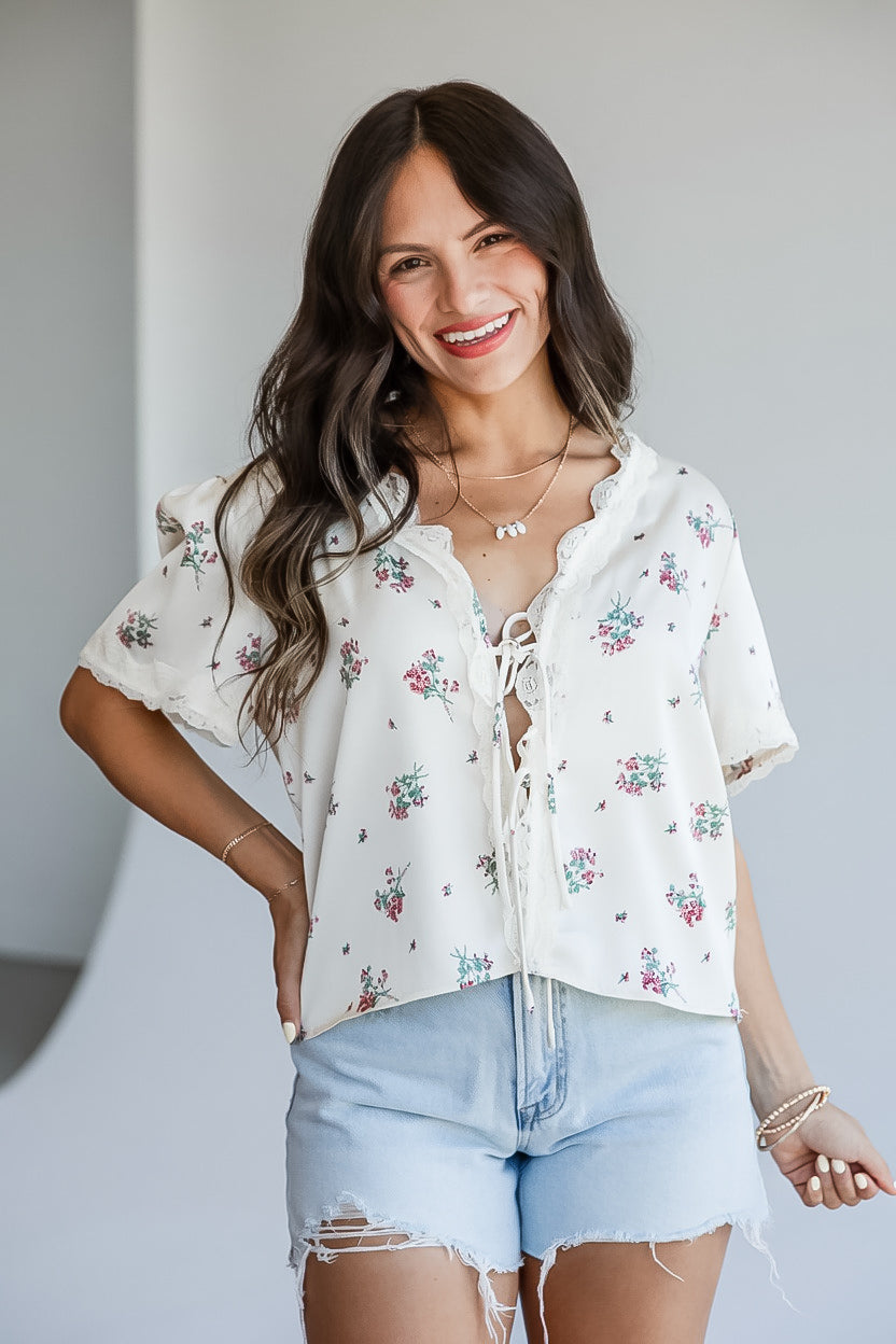 A woman modeling a white short-sleeve blouse with a delicate pink and green floral print, lace-up front, and ruffled collar, paired with light-wash denim shorts.