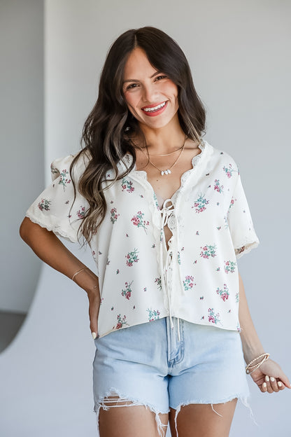 A woman modeling a white short-sleeve blouse with a delicate pink and green floral print, lace-up front, and ruffled collar, paired with light-wash denim shorts.