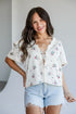 A woman modeling a white short-sleeve blouse with a delicate pink and green floral print, lace-up front, and ruffled collar, paired with light-wash denim shorts.