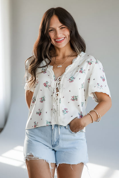 A woman wearing a short-sleeve cream blouse with a delicate pink and green floral print, lace-up front, and ruffled trim, paired with light-wash denim shorts.
