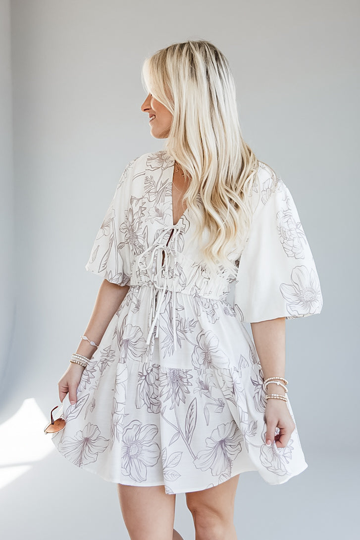 A woman wearing a white tiered mini dress with a gray floral line print, shown from the back-three-quarter view against a light gray studio background.