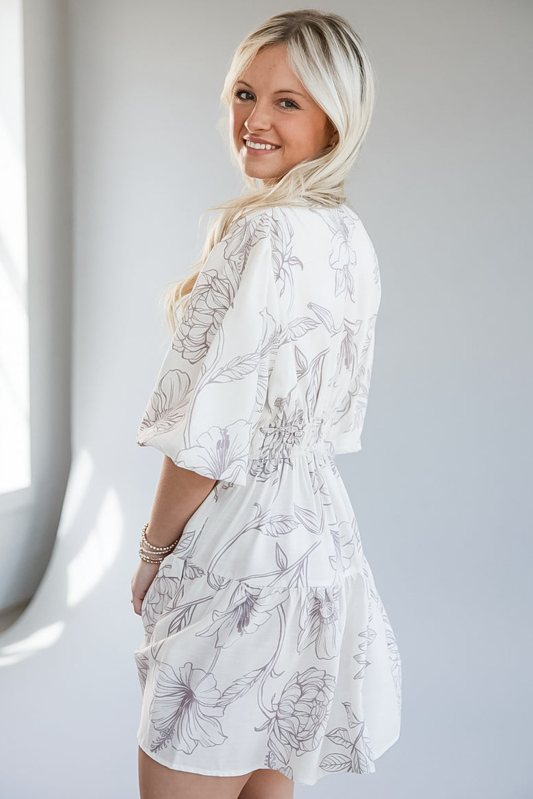 A woman wearing a white short-sleeve dress with a gray floral line-art print, photographed in a bright indoor setting.