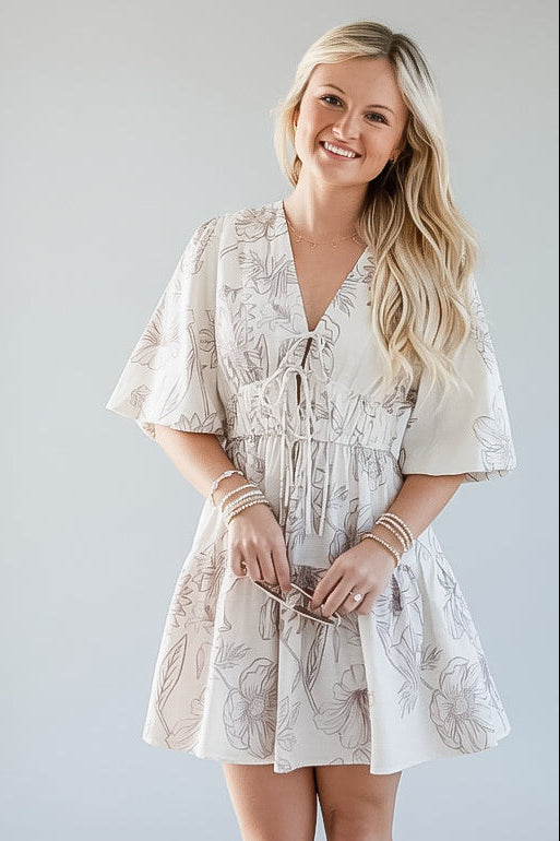 A woman modeling a short cream-colored dress with a light brown floral pattern, featuring a tie-front waist and flutter sleeves, set against a plain light gray studio background.