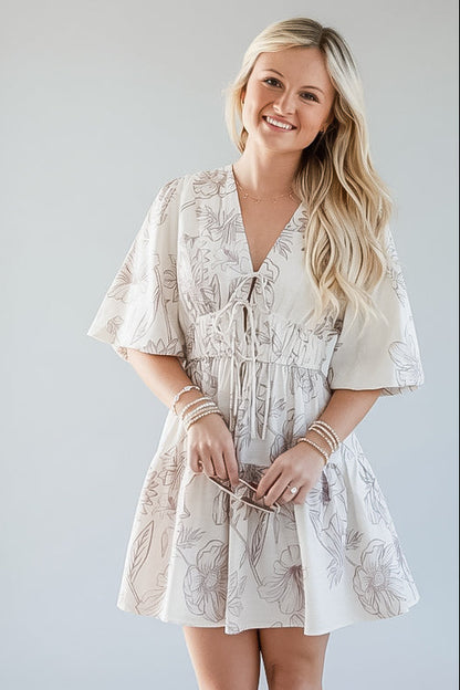 A woman modeling a short cream-colored dress with a light brown floral pattern, featuring a tie-front waist and flutter sleeves, set against a plain light gray studio background.