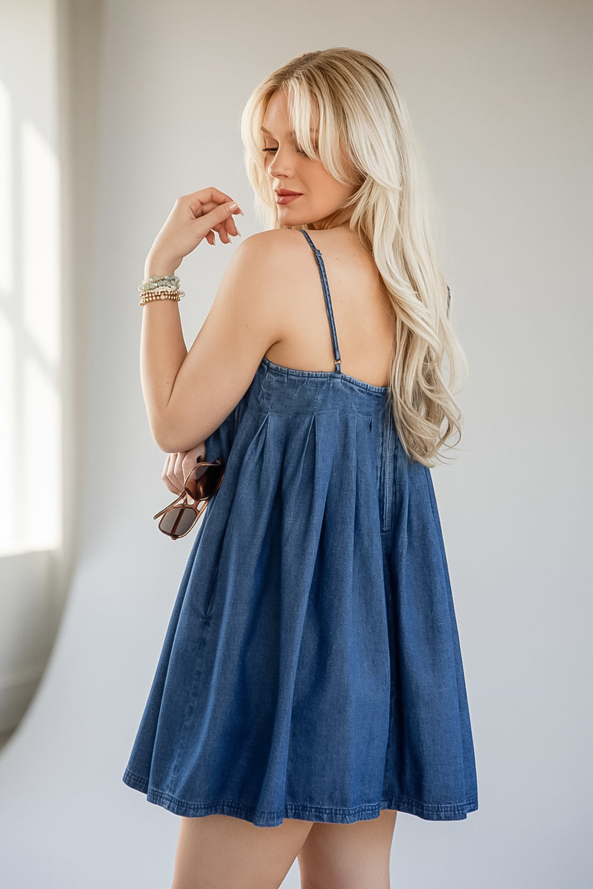 A blonde woman wearing a blue denim mini dress, photographed from behind in a bright indoor setting with a light gray wall background.
