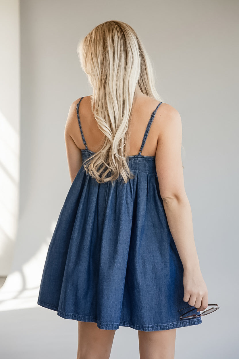 A woman with long blonde hair is shown from behind wearing a blue denim slip dress with thin straps and a gathered waist, standing against a light gray wall.