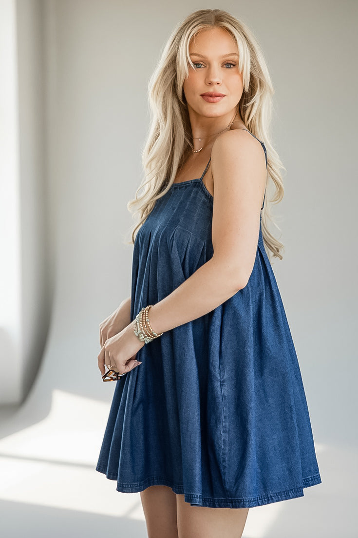 A blonde woman wearing a blue denim camisole dress with a flared skirt, standing in a brightly lit indoor setting.