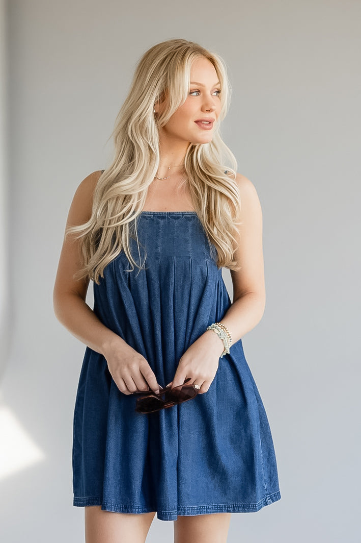 A blonde woman wearing a blue denim strapless dress, holding a pair of sunglasses against a plain light gray background.