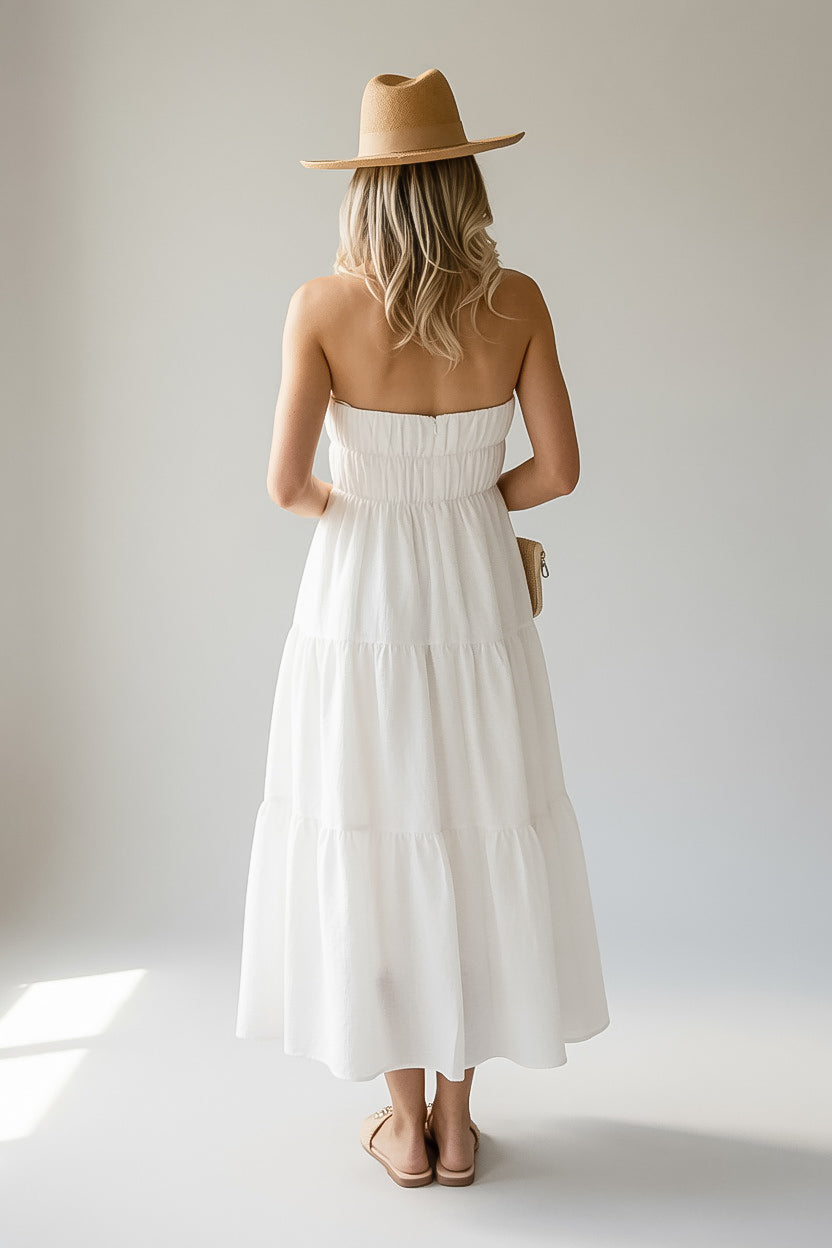 A woman stands with her back to the camera wearing a strapless white tiered maxi dress, a tan wide-brim hat, and tan sandals against a plain light gray studio background.