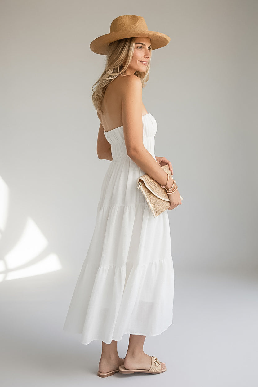 A woman wearing a strapless white tiered maxi dress, a tan wide-brim hat, and beige flat sandals, holding a woven clutch against a light gray studio background.