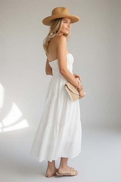 A woman wearing a strapless white tiered maxi dress, a tan wide-brim hat, and beige flat sandals, holding a woven clutch against a light gray studio background.