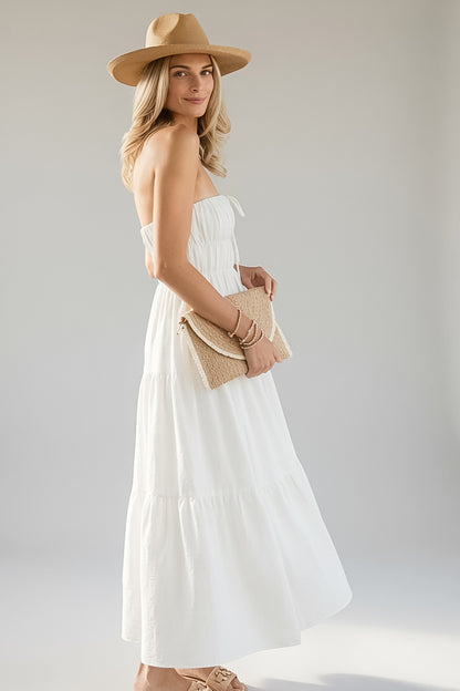 A woman modeling a strapless white tiered maxi dress, styled with a tan wide-brim hat, a woven tan clutch, and tan sandals against a light gray studio background.