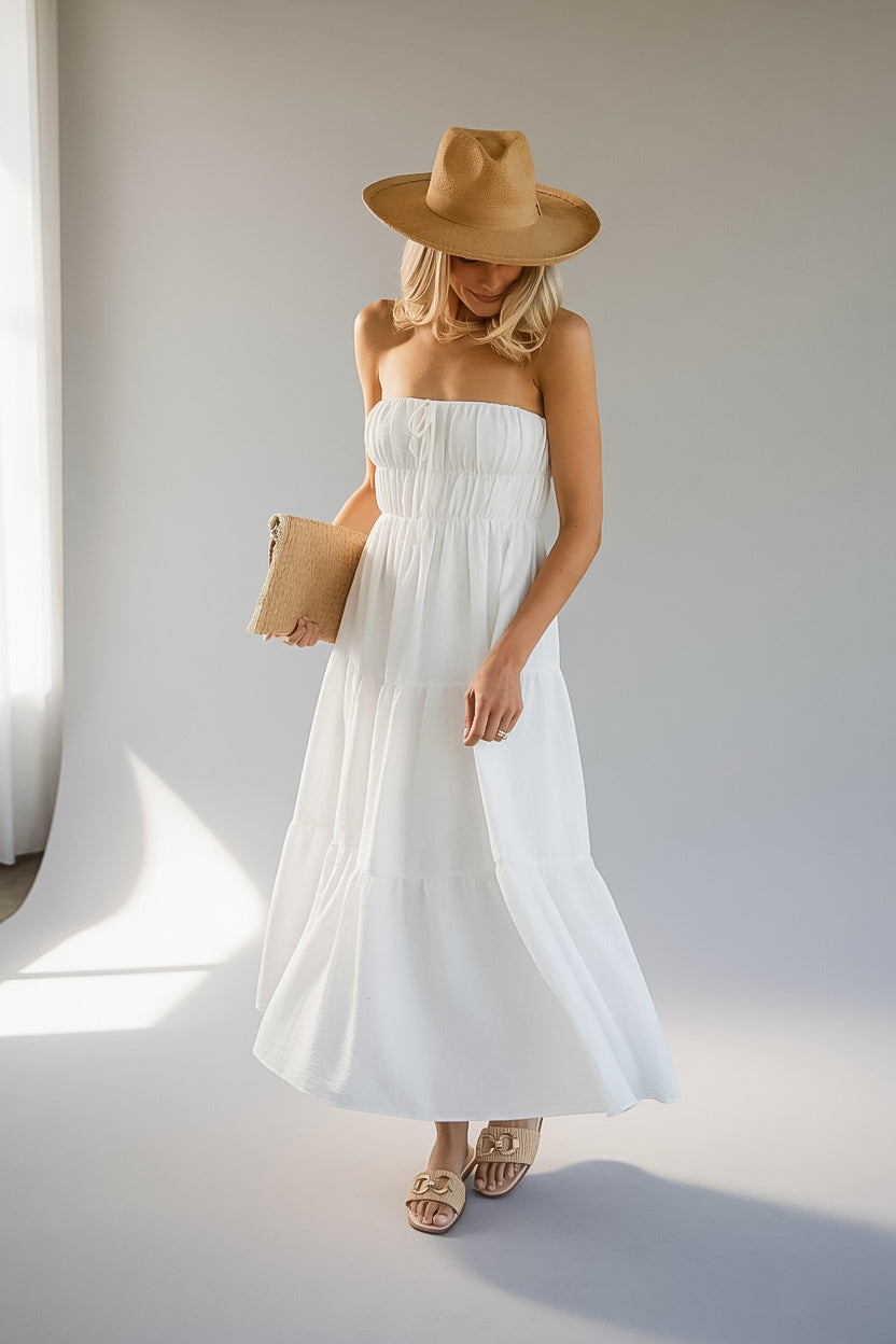 A woman modeling a white strapless tiered maxi dress, styled with a tan wide-brim hat, a beige clutch, and tan sandals, standing in a brightly lit studio.