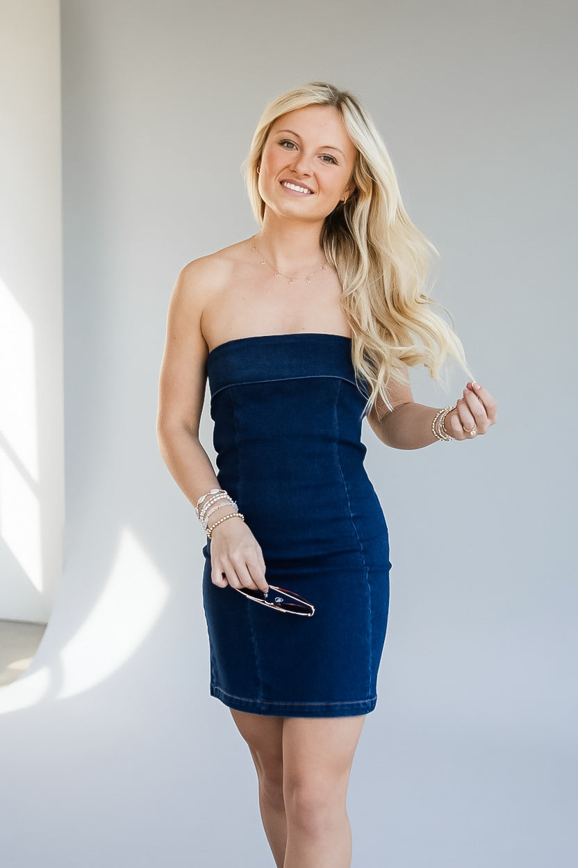 A blonde woman wearing a strapless dark blue denim mini dress, holding sunglasses, standing against a light gray wall with soft window light.