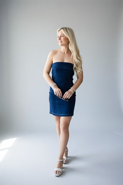 A blonde woman models a strapless dark blue mini dress with a textured, denim-like finish, styled with white heeled sandals and bracelets, against a light gray studio background.