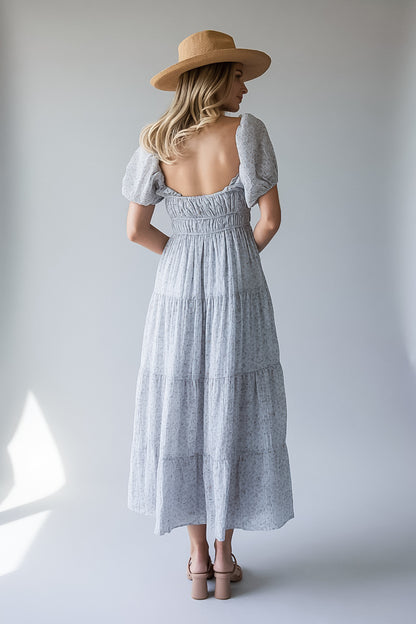 A woman viewed from behind wearing a light gray and white patterned tiered maxi dress with puff sleeves and a smocked back, paired with a tan wide-brim hat and beige heels, set against a plain light gray studio background.