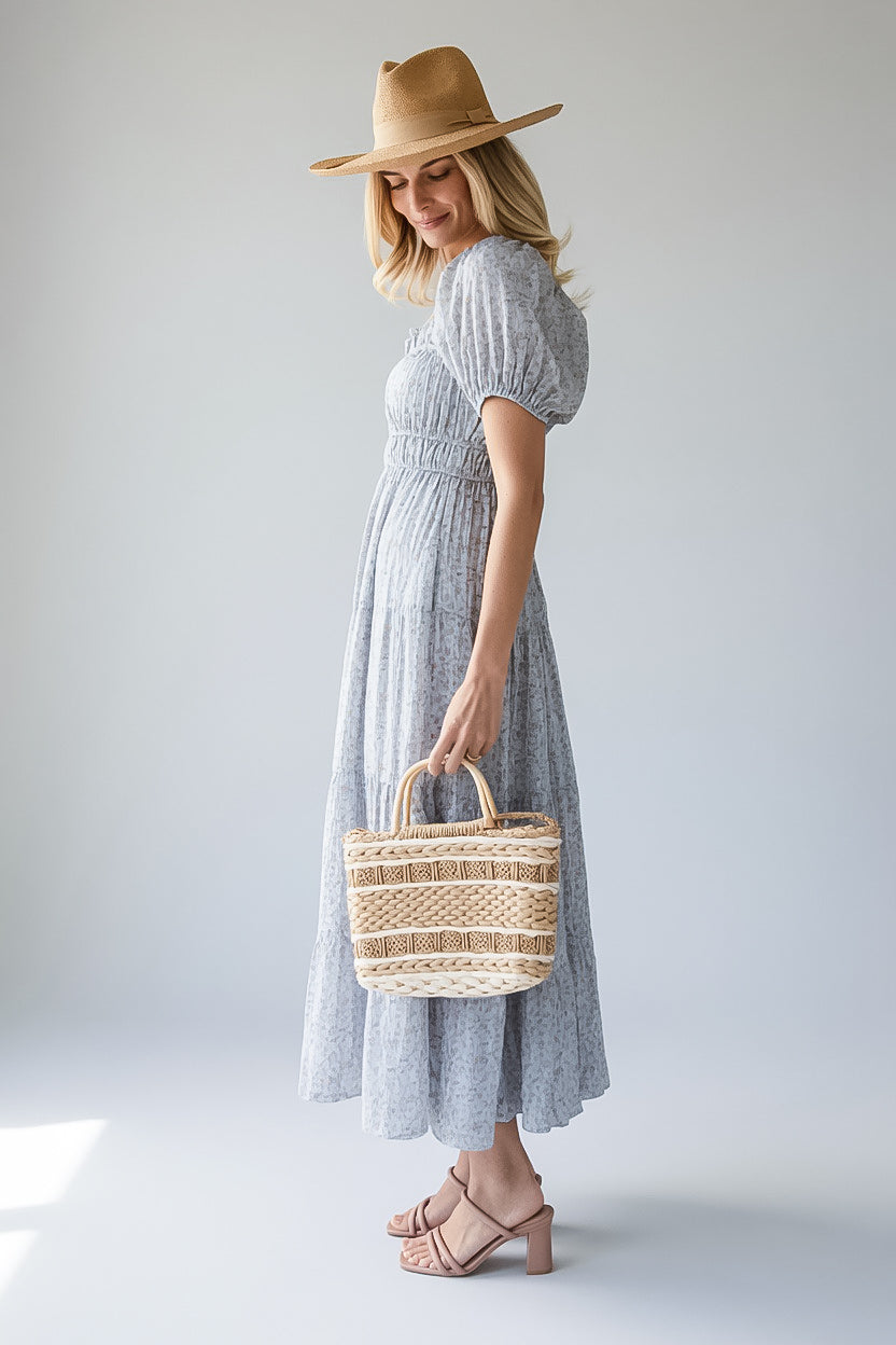 A woman wearing a light blue-gray textured maxi dress with puff sleeves, a tan wide-brim hat, and holding a woven straw tote bag, standing against a light gray studio background.