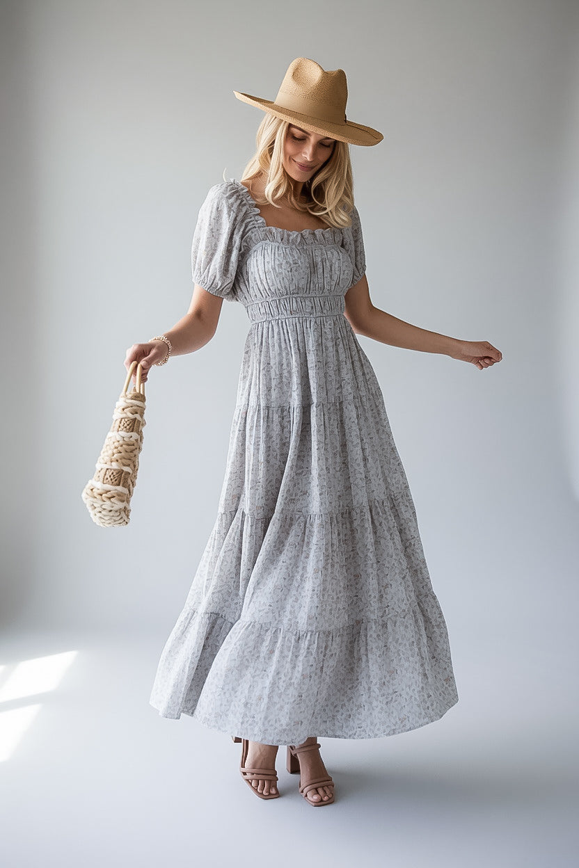 A woman modeling a light blue and white floral print tiered maxi dress with puff sleeves, a smocked bodice, and a square neckline. She is wearing a tan wide-brim hat and holding a woven straw bag against a neutral light gray studio background.