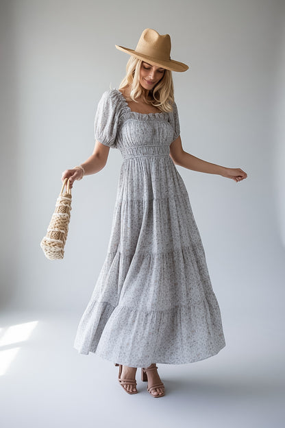 A woman modeling a light blue and white floral print tiered maxi dress with puff sleeves, a smocked bodice, and a square neckline. She is wearing a tan wide-brim hat and holding a woven straw bag against a neutral light gray studio background.