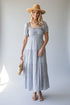 A woman modeling a light blue and white textured tiered midi dress with puff sleeves and a smocked bodice, styled with a wide-brimmed straw hat and a woven clutch bag against a light gray studio background.
