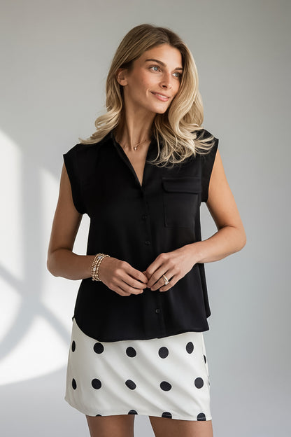 A woman modeling a sleeveless black button-front blouse with a collar and chest pocket, paired with a white skirt featuring large black polka dots, against a light gray studio background.