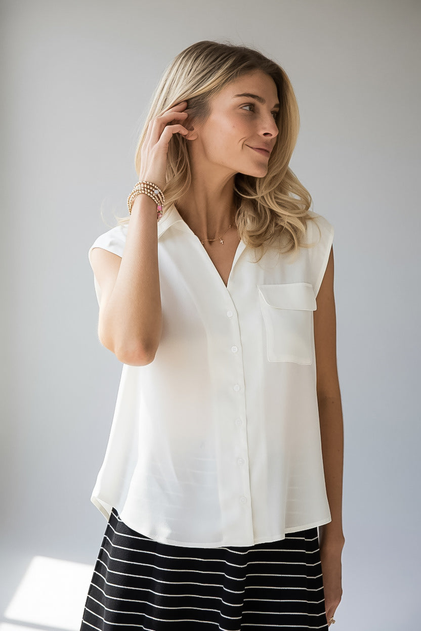 A woman wearing a sleeveless white button-up blouse with a chest pocket, paired with a black and white striped skirt, standing against a light gray background.
